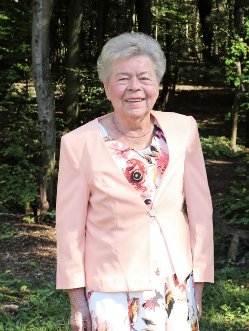 L&auml;chelnde &auml;ltere Frau in einem bl&uuml;tenmusternden Kleid und einer rosa Jacke vor einem Waldhintergrund.