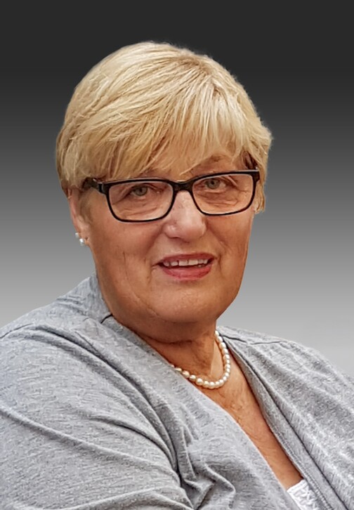 Porträt einer Frau mit blonden, kurzen Haaren und Brille, die in einem grauen Oberteil und Perlenkette lächelt.