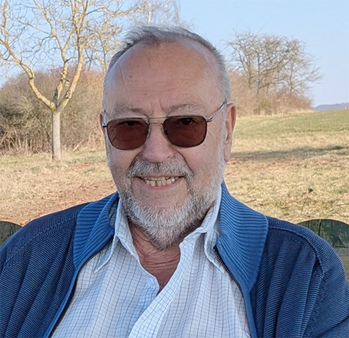 Lächelnder älterer Mann mit Brille und Bart, sitzend vor einer freien Landschaft mit Bäumen und blauem Himmel.