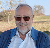 L&auml;chelnder &auml;lterer Mann mit Brille und Bart, sitzend vor einer freien Landschaft mit B&auml;umen und blauem Himmel.