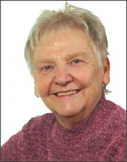 Porträt einer älteren Frau mit kurzen grauen Haaren und Pullover, lächelnd vor hellem Hintergrund.