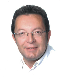 Portr&auml;t eines Mannes mit Brille, der l&auml;chelt, vor einem wei&szlig;en Hintergrund. Er tr&auml;gt ein helles Oberteil.