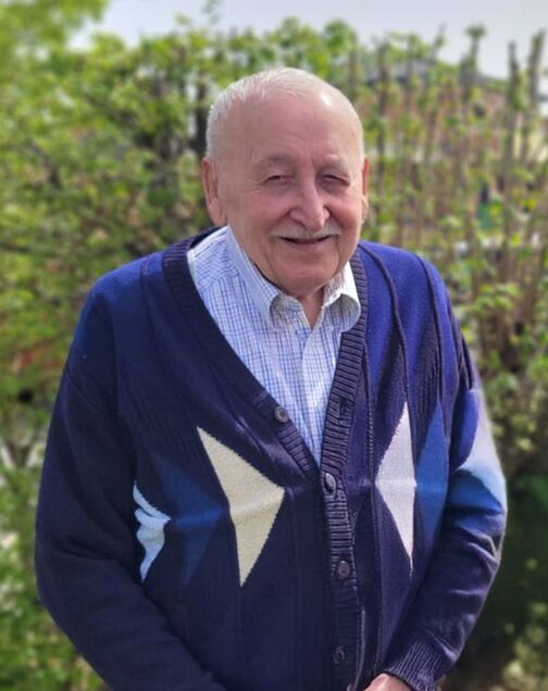 Älterer Mann in einem gemusterten Pullover lächelt vor grünem Hintergrund im Freien.