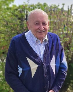 Älterer Mann in einem gemusterten Pullover lächelt vor grünem Hintergrund im Freien.