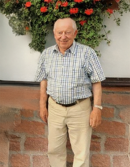 &Auml;lterer Mann mit Karohemd und beigefarbenen Hosen steht vor einer Wand mit Blumenampeln.