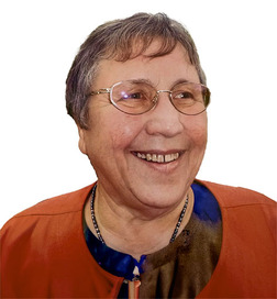 L&auml;chelnde Frau mit Brille und kurzem Haar, tr&auml;gt einen orangefarbenen Blazer und sitzt vor einem wei&szlig;en Hintergrund.