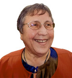 Lächelnde Frau mit Brille und kurzem Haar, trägt einen orangefarbenen Blazer und sitzt vor einem weißen Hintergrund.