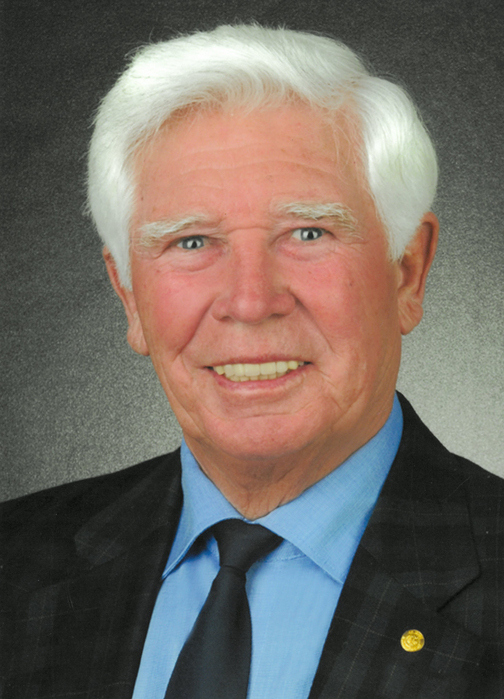 Portrait eines &auml;lteren Mannes mit wei&szlig;em Haar, blauer Hemd und schwarzem Anzug, l&auml;chelnd vor neutralem Hintergrund.