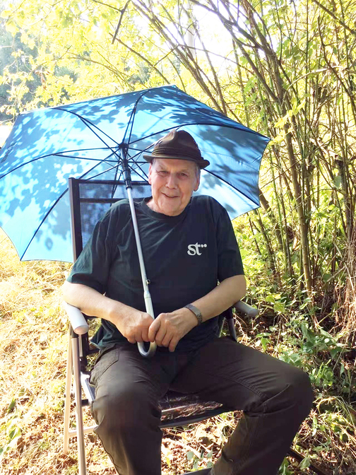 &Auml;lterer Mann sitzt entspannt auf einem Stuhl, gehalten von einem blauen Sonnenschirm in einer nat&uuml;rlichen Umgebung.