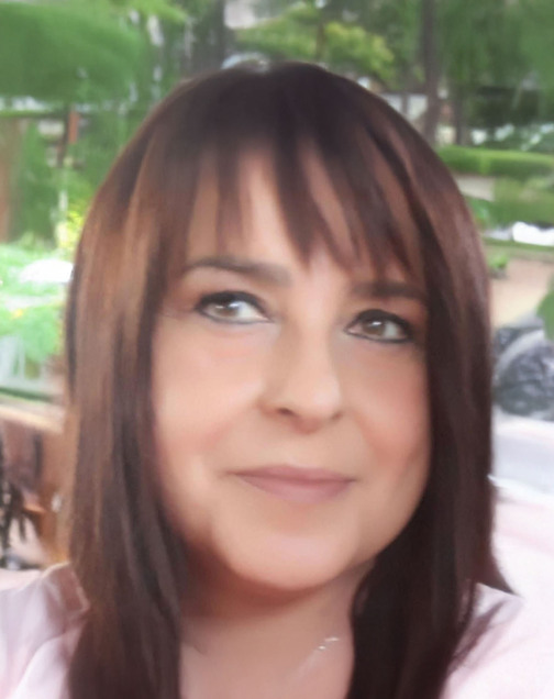 Nahaufnahme einer Person mit langen, braunen Haaren und einem neutralen Ausdruck, im Hintergrund Pflanzen und verschwommenes Ambiente.