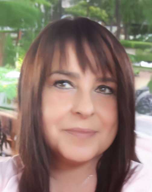 Nahaufnahme einer Person mit langen, braunen Haaren und einem neutralen Ausdruck, im Hintergrund Pflanzen und verschwommenes Ambiente.