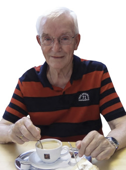 &Auml;lterer Mann mit Brille, der in einer Tasse Kaffee r&uuml;hrt, sitzt an einem Tisch mit einer Kuchenteller.