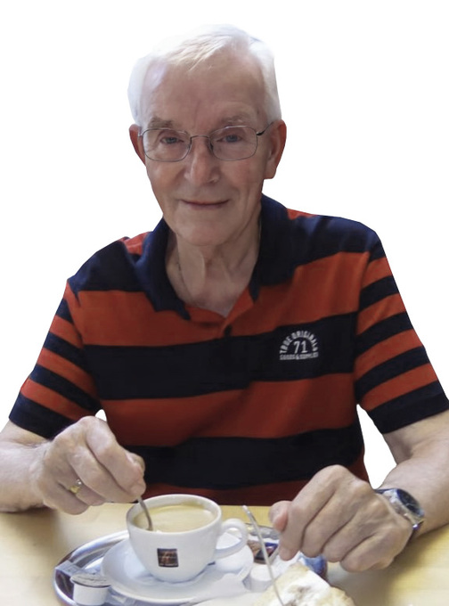 &Auml;lterer Mann mit Brille, der in einer Tasse Kaffee r&uuml;hrt, sitzt an einem Tisch mit einer Kuchenteller.