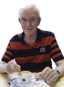 &Auml;lterer Mann mit Brille, der in einer Tasse Kaffee r&uuml;hrt, sitzt an einem Tisch mit einer Kuchenteller.