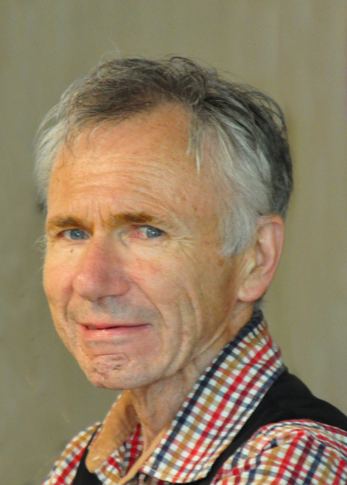 Portr&auml;t eines &auml;lteren Mannes mit grauem Haar und schottischem Karomusterhemd, l&auml;chelnd vor neutralem Hintergrund.