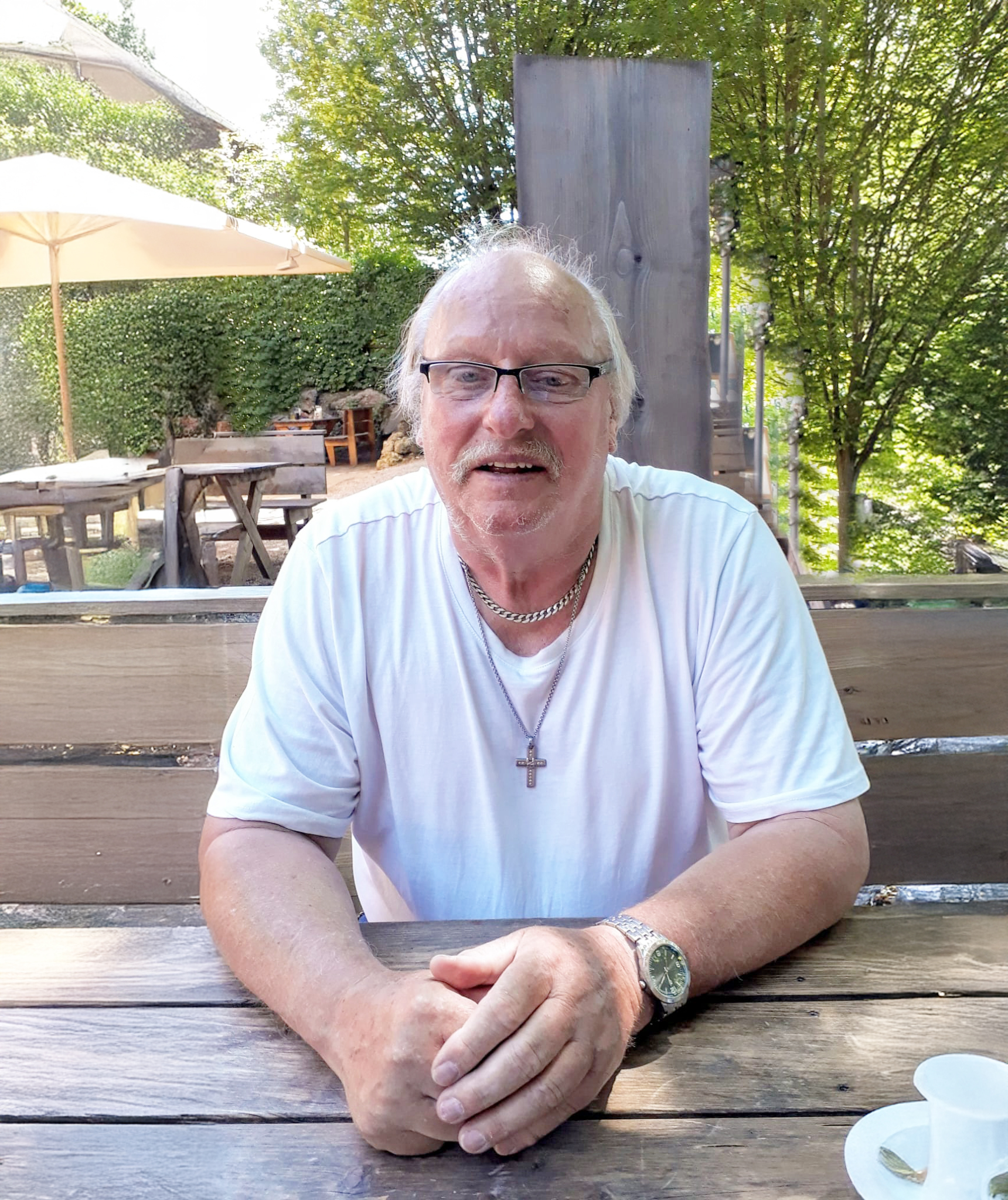 Mann mit Brille und Kreuzanhänger sitzt an einem Holztisch im Freien, umgeben von Bäumen und Sonnenschirmen.