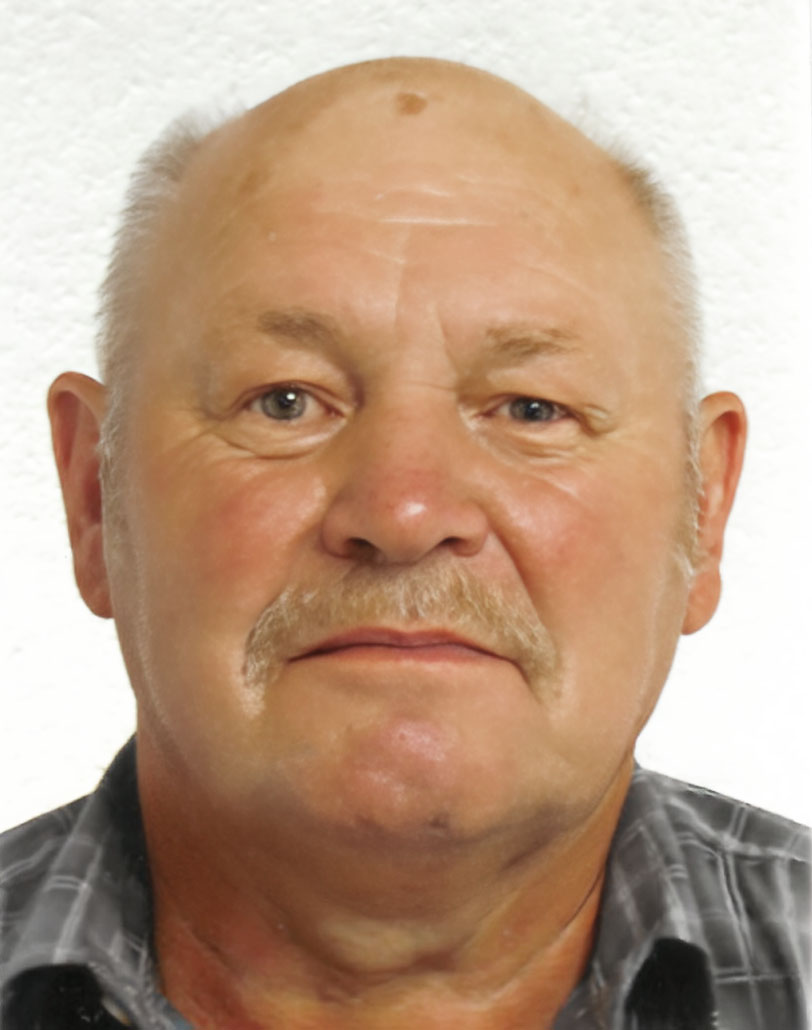 Porträt eines älteren Mannes mit kurzem, grauem Haar und Schnurrbart, vor einem weißen Hintergrund.