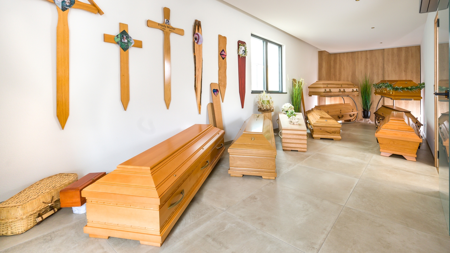 Raum mit mehreren Holzkreuzen an der Wand und verschiedenen hölzernen Särgen, einige dekoriert mit Pflanzen.