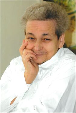 &Auml;ltere Frau mit kurzen grauen Haaren, in wei&szlig;em Oberteil, l&auml;chelt nachdenklich mit der Hand am Gesicht.