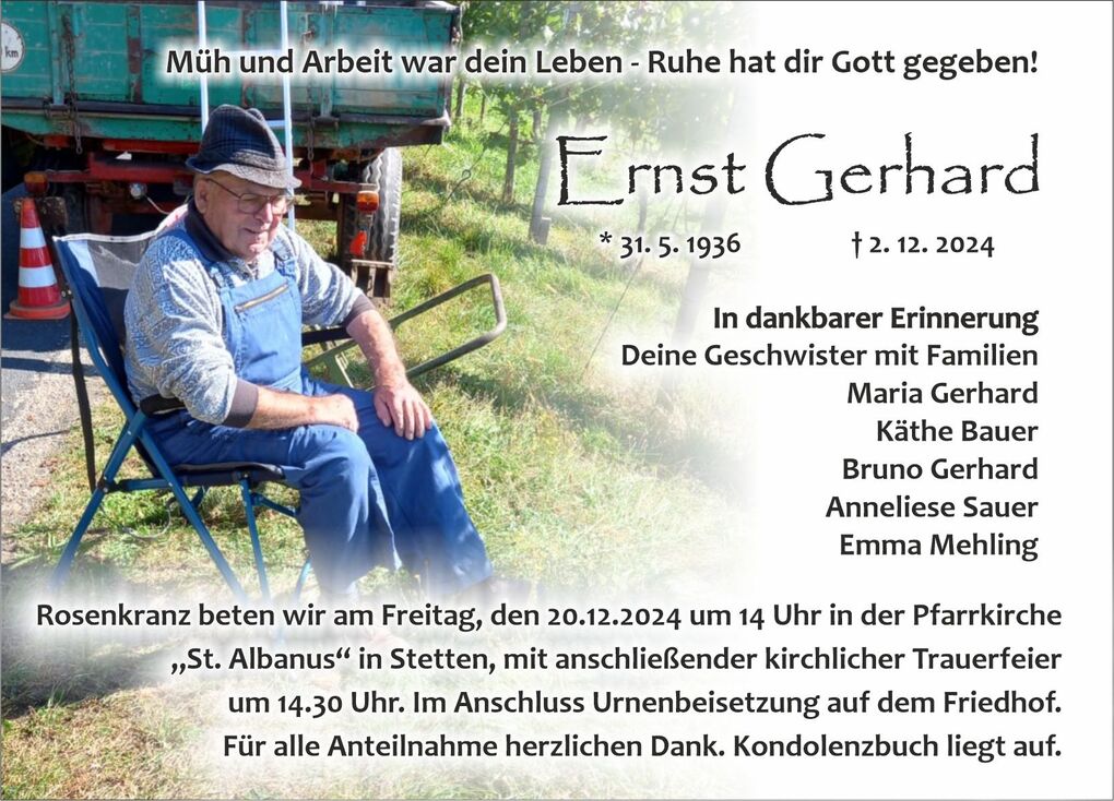 Ein &auml;lterer Mann in Arbeitskleidung sitzt auf einem Stuhl im Freien, umgeben von Pflanzen und einem gr&uuml;nen Anh&auml;nger.