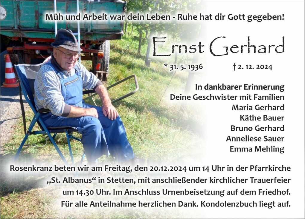 Ein &auml;lterer Mann in Arbeitskleidung sitzt auf einem Stuhl im Freien, umgeben von Pflanzen und einem gr&uuml;nen Anh&auml;nger.