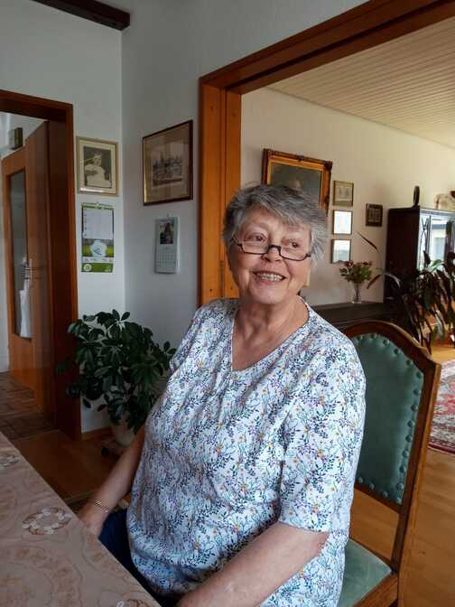 Lächelnde ältere Dame mit grau-blonden Haaren sitzt an einem Tisch in einem gemütlichen Wohnraum.