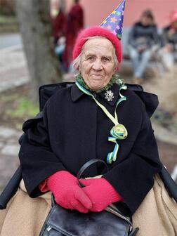 &Auml;ltere Frau mit rotem Hut und Handschuhen, l&auml;chelt in einem Rollstuhl, umgeben von einer festlichen Atmosph&auml;re.