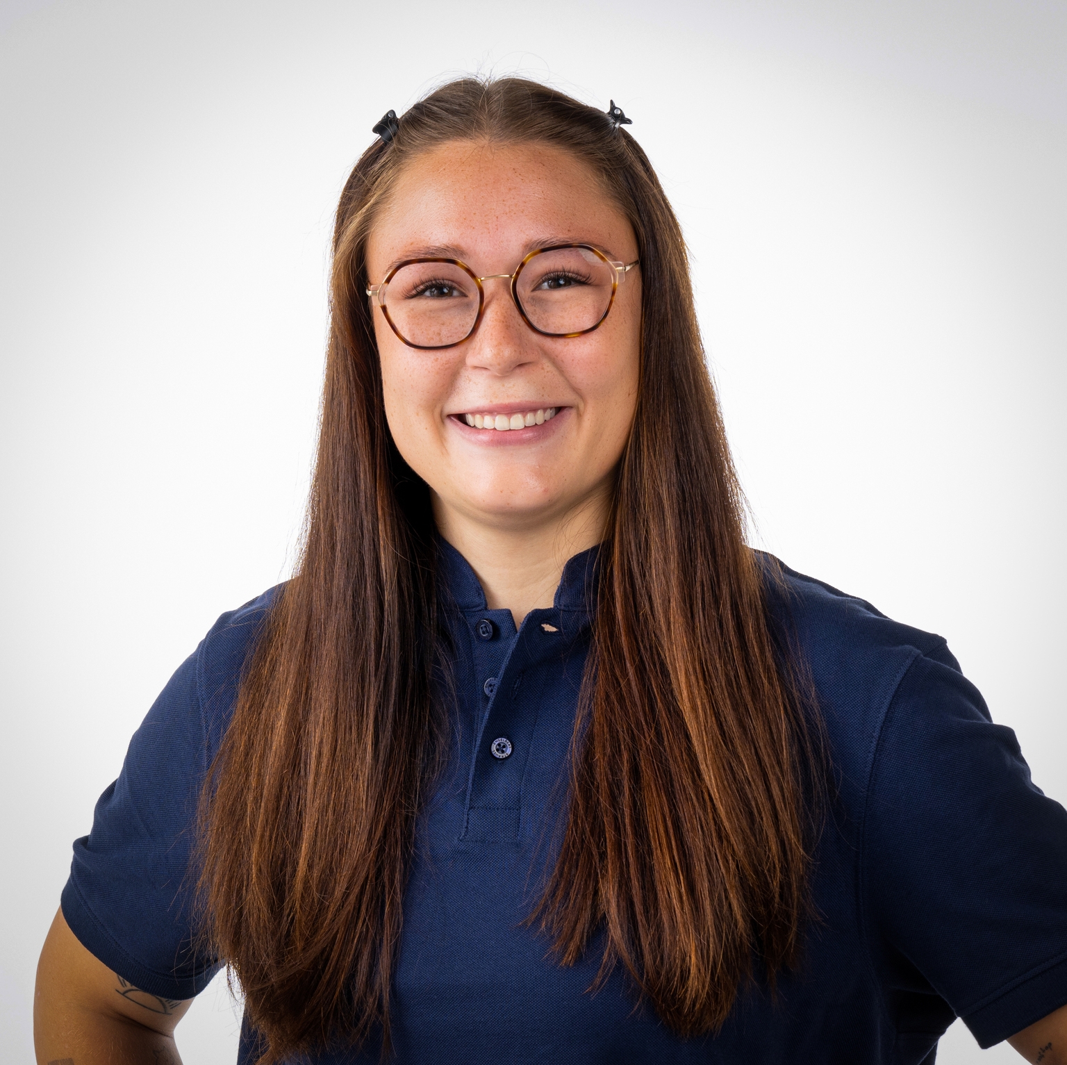 Lächelnde junge Frau mit langen, brünetten Haaren und Brille in dunkelblauem Polohemd vor hellem Hintergrund.