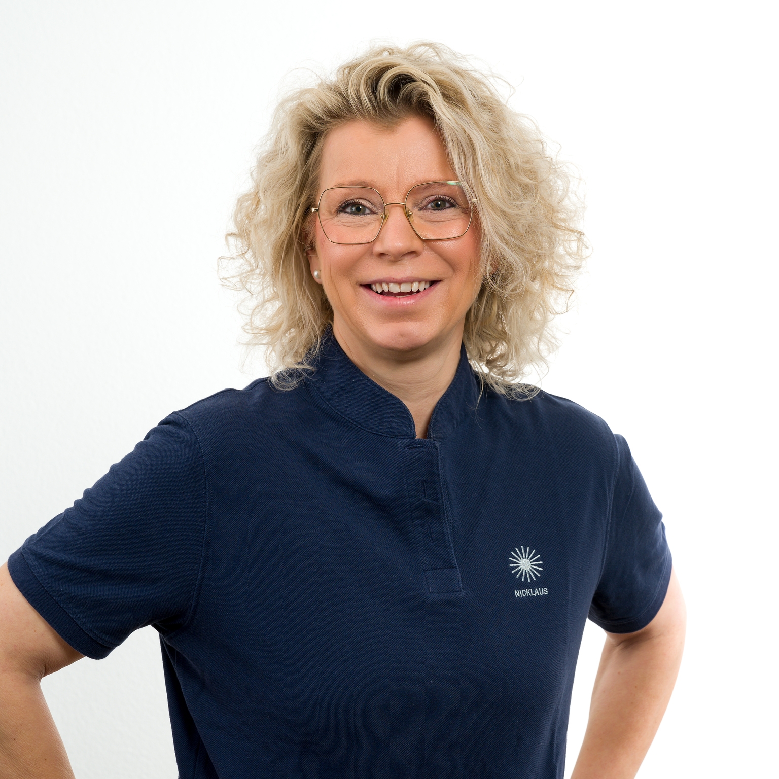 Lächelnde Frau mit lockigem Haar in einem blauen Shirt vor einem neutralen Hintergrund. Sie hat eine Brille und steht mit verschränkten Armen.