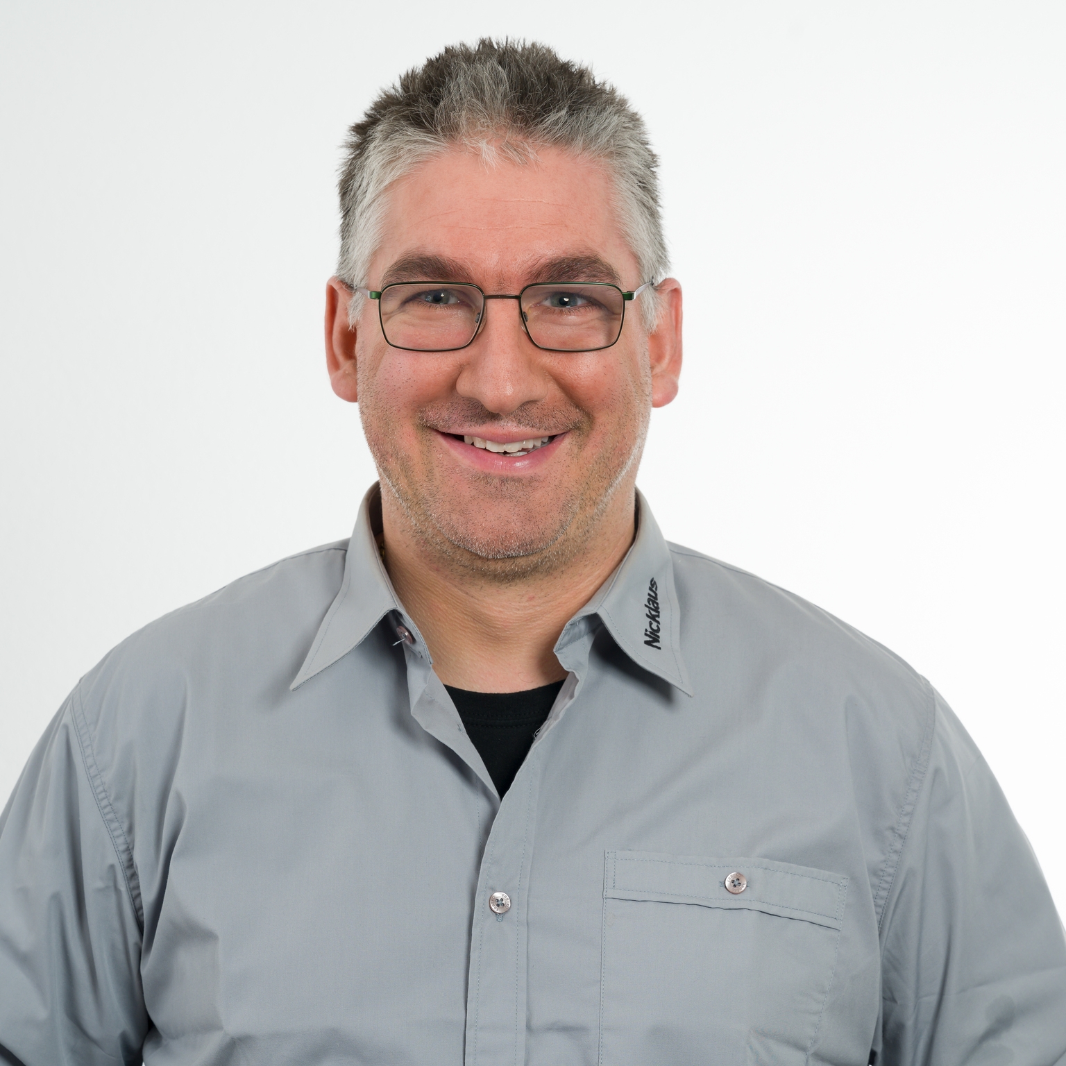 Mann mit Brille und grauem Hemd lächelt freundlich vor hellem Hintergrund.