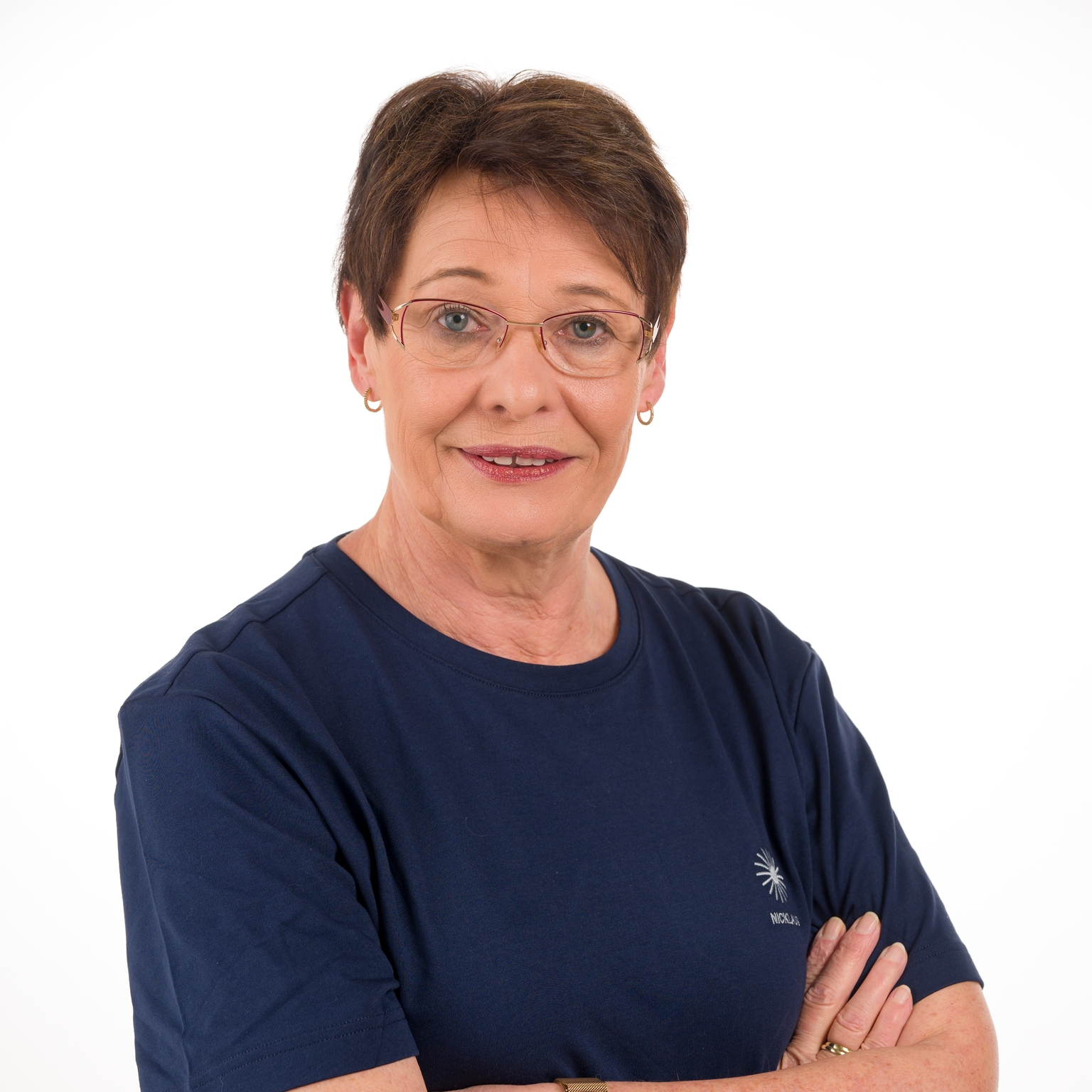 Porträt einer lächelnden Frau mit kurzen Haaren, Brille und blauer T-Shirt, die die Arme vor der Brust verschränkt.