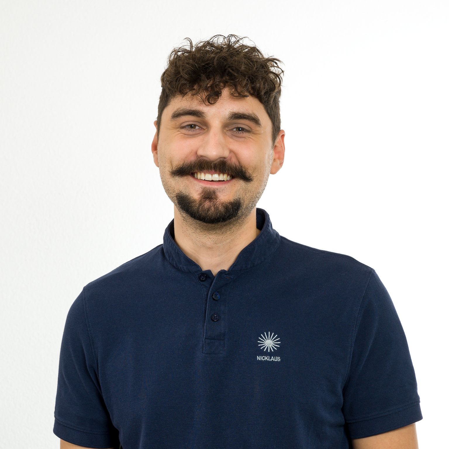 Porträt eines lächelnden Mannes mit lockigem Haar und Schnurrbart, der ein dunkelblaues Poloshirt trägt. Hintergründe sind weiß.