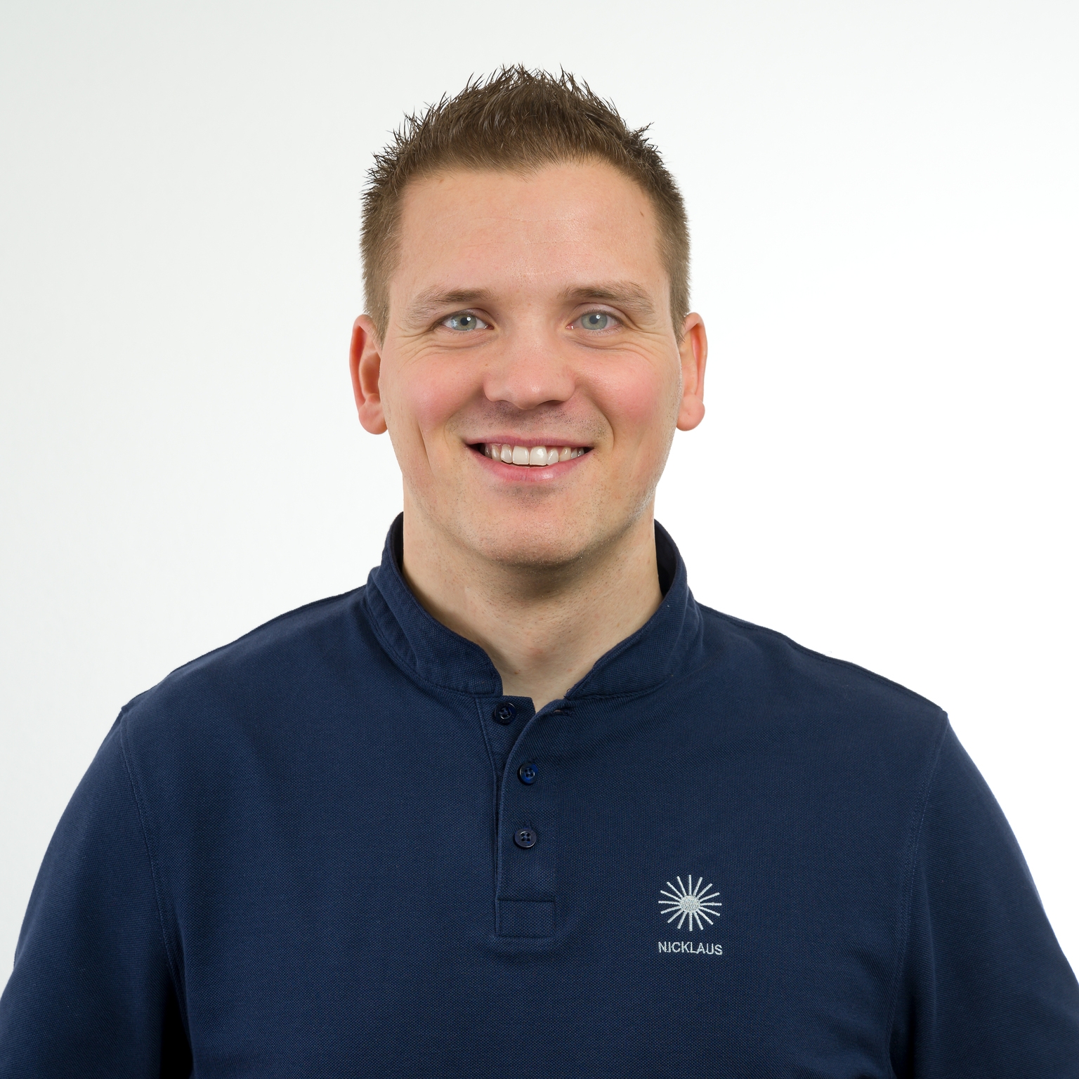 Porträt eines lächelnden Mannes mit kurzem, straffem Haar, der ein dunkles Poloshirt trägt, vor hellem Hintergrund.