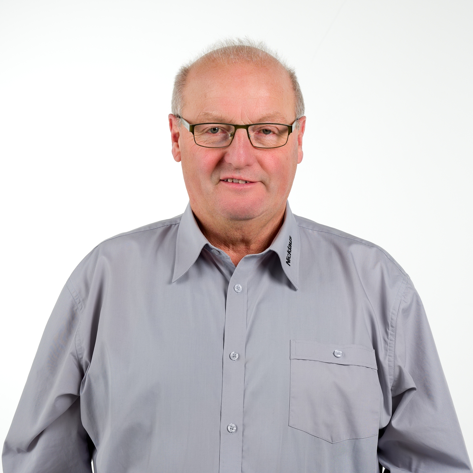 Porträt eines Mannes mit Brille, grauhem Hemd und kurzen Haaren, vor einem weißen Hintergrund.