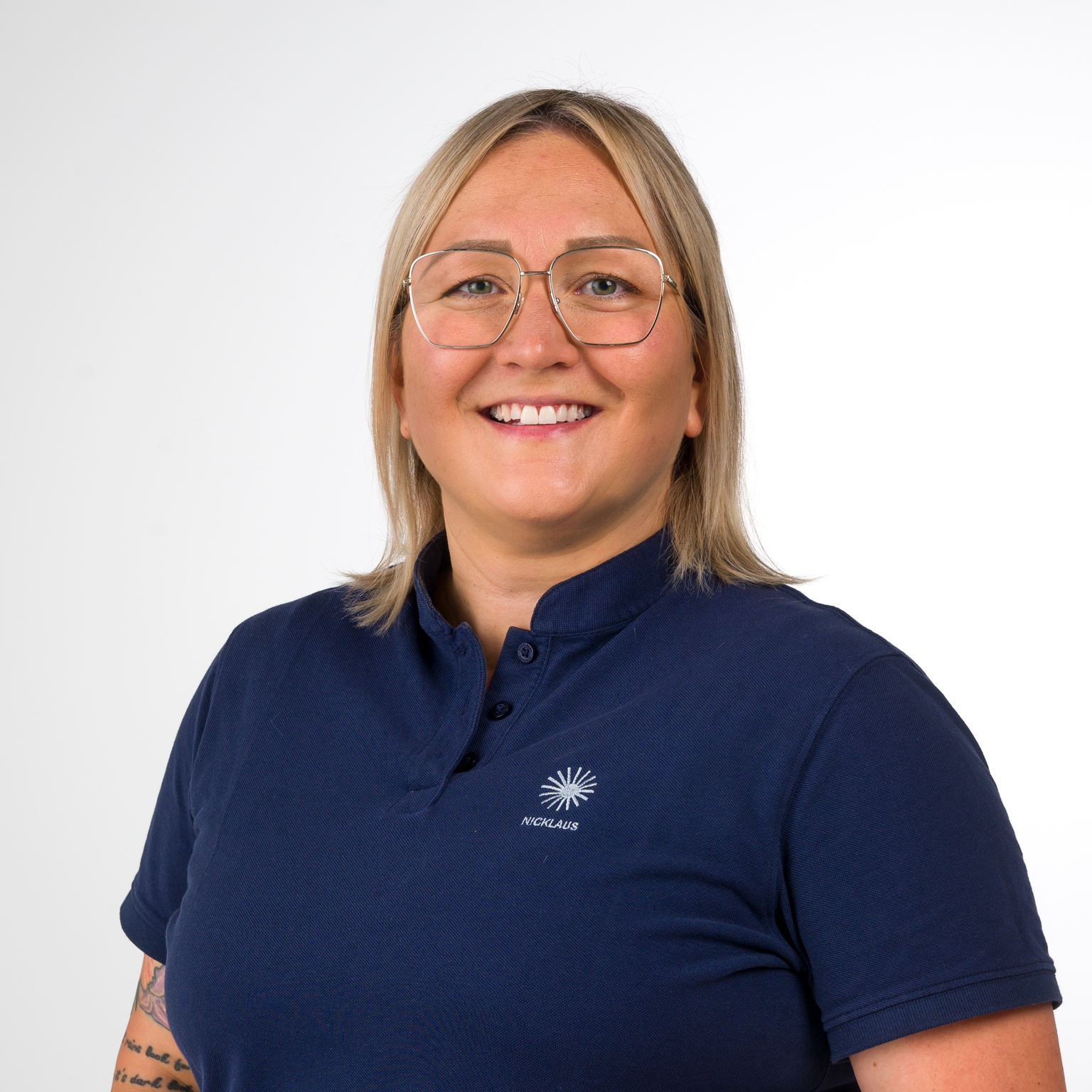 Portrait einer lächelnden Frau mit kurzen blonden Haaren, Brille und dunkelblauem Polohemd. Hintergrund ist hell.