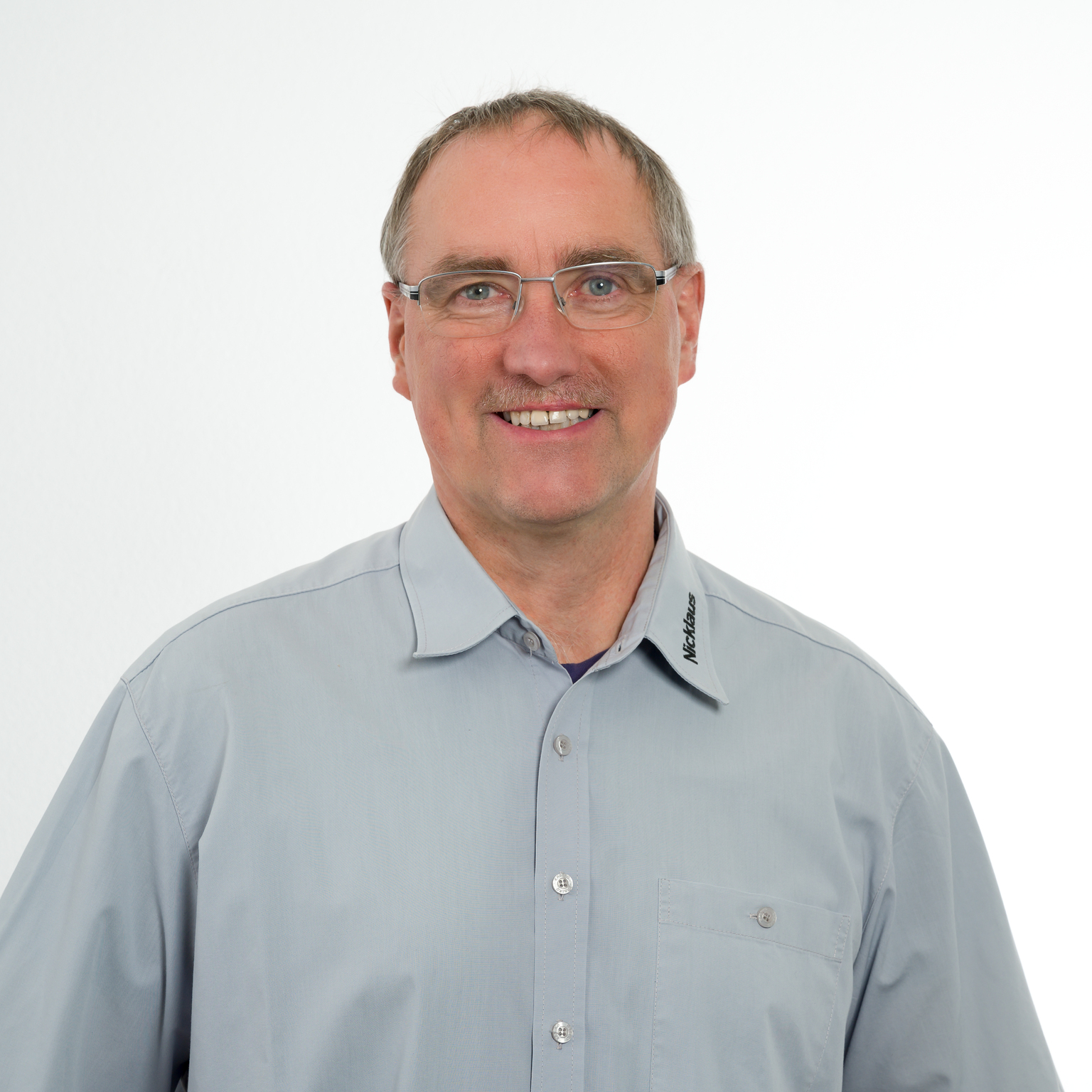 Portr&auml;t eines l&auml;chelnden Mannes mit Brille in einem grauen Hemd vor einem wei&szlig;en Hintergrund.