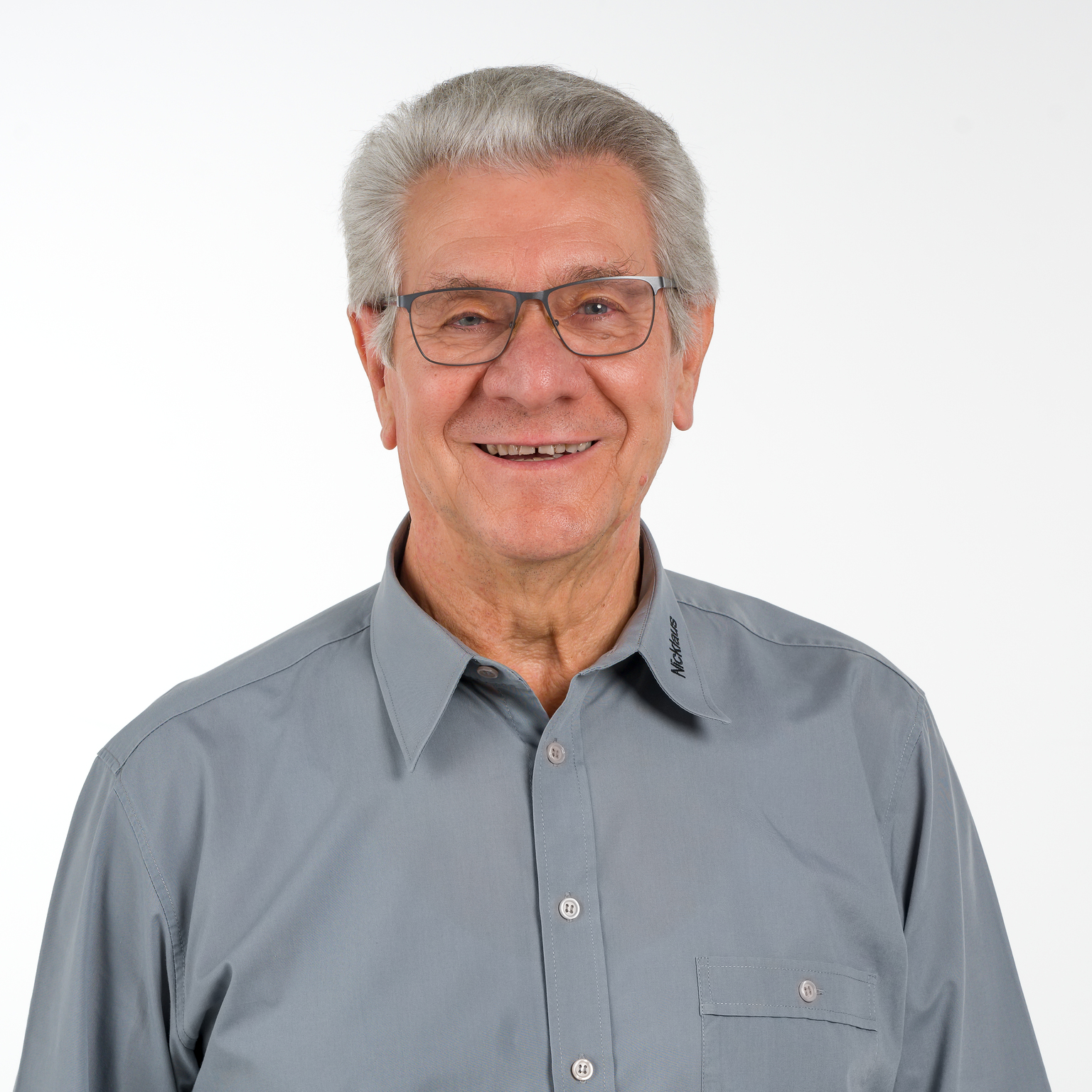 &Auml;lterer Mann mit Brille und grauem Hemd l&auml;chelt vor neutralem Hintergrund.