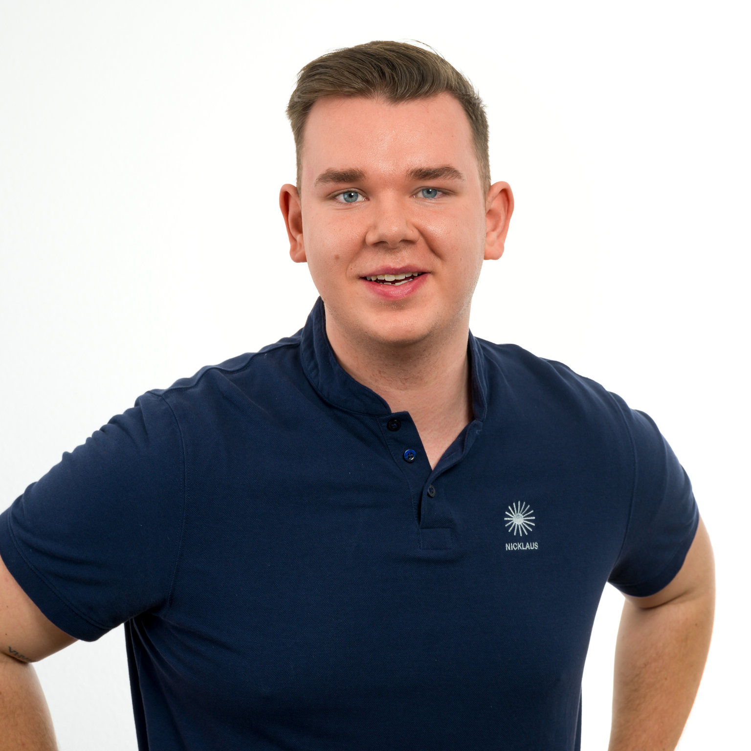 Junger Mann mit kurzem Haar und blauem Poloshirt l&auml;chelt freundlich vor einem wei&szlig;en Hintergrund.
