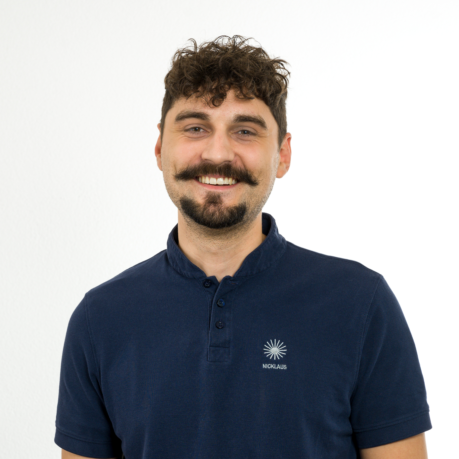 Portr&auml;t eines l&auml;chelnden Mannes mit lockigem Haar und Schnurrbart, der ein dunkelblaues Poloshirt tr&auml;gt. Hintergr&uuml;nde sind wei&szlig;.