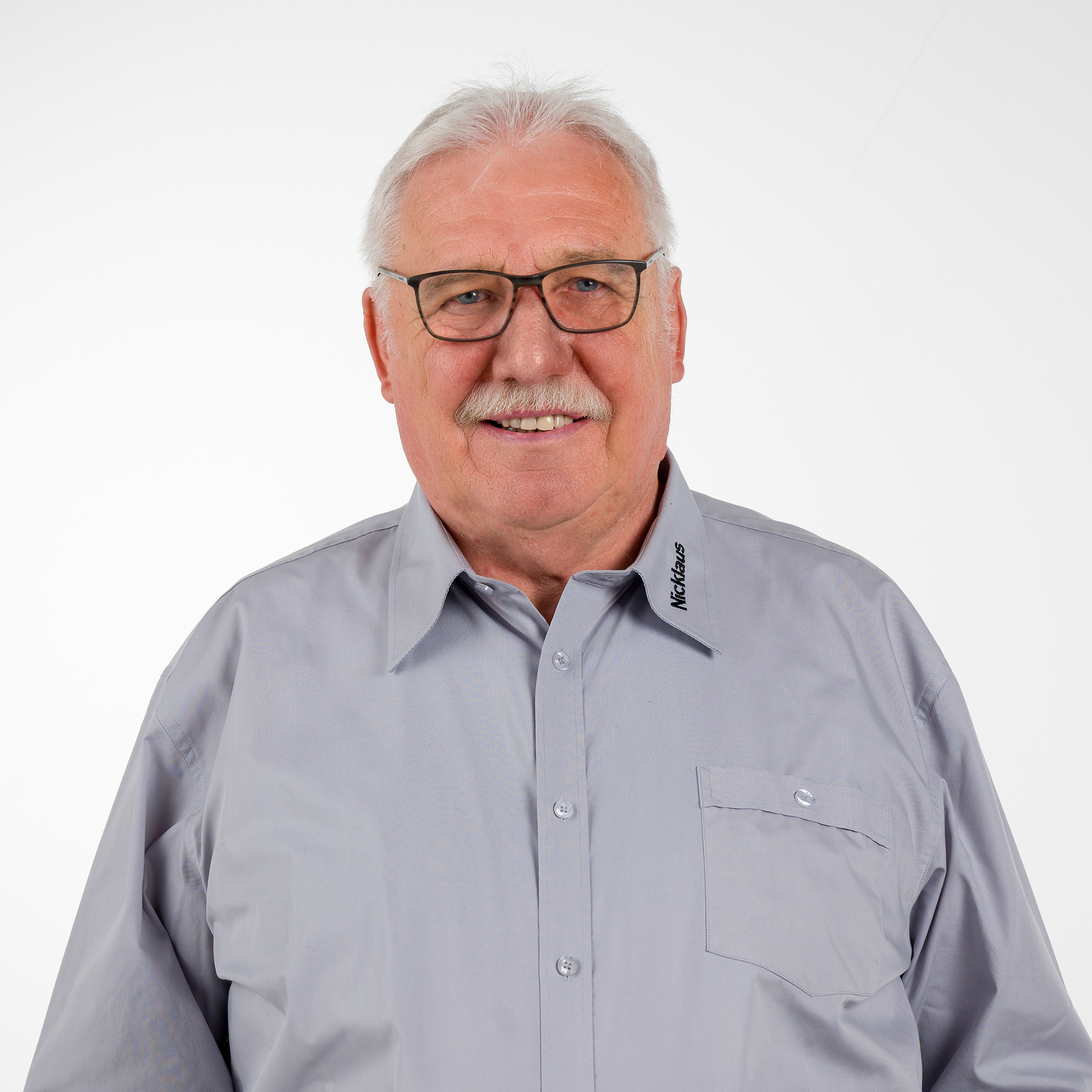 Portrait eines l&auml;chelnden Mannes mit Brille in einem grauen Hemd vor wei&szlig;em Hintergrund.