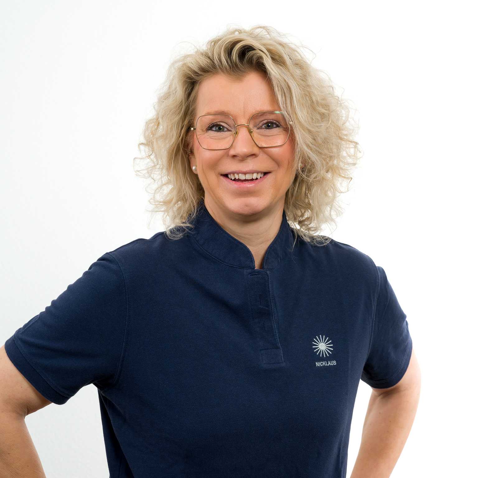 L&auml;chelnde Frau mit lockigem Haar in einem blauen Shirt vor einem neutralen Hintergrund. Sie hat eine Brille und steht mit verschr&auml;nkten Armen.