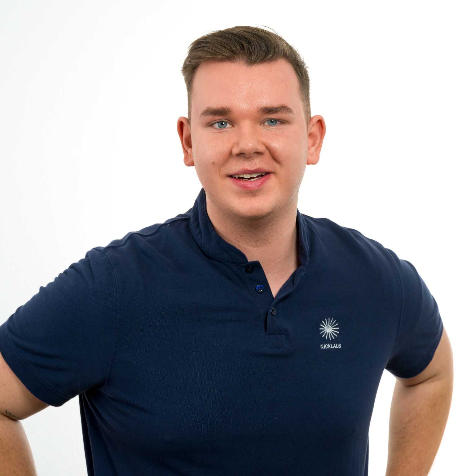Junger Mann mit kurzem Haar und blauem Poloshirt l&auml;chelt freundlich vor einem wei&szlig;en Hintergrund.