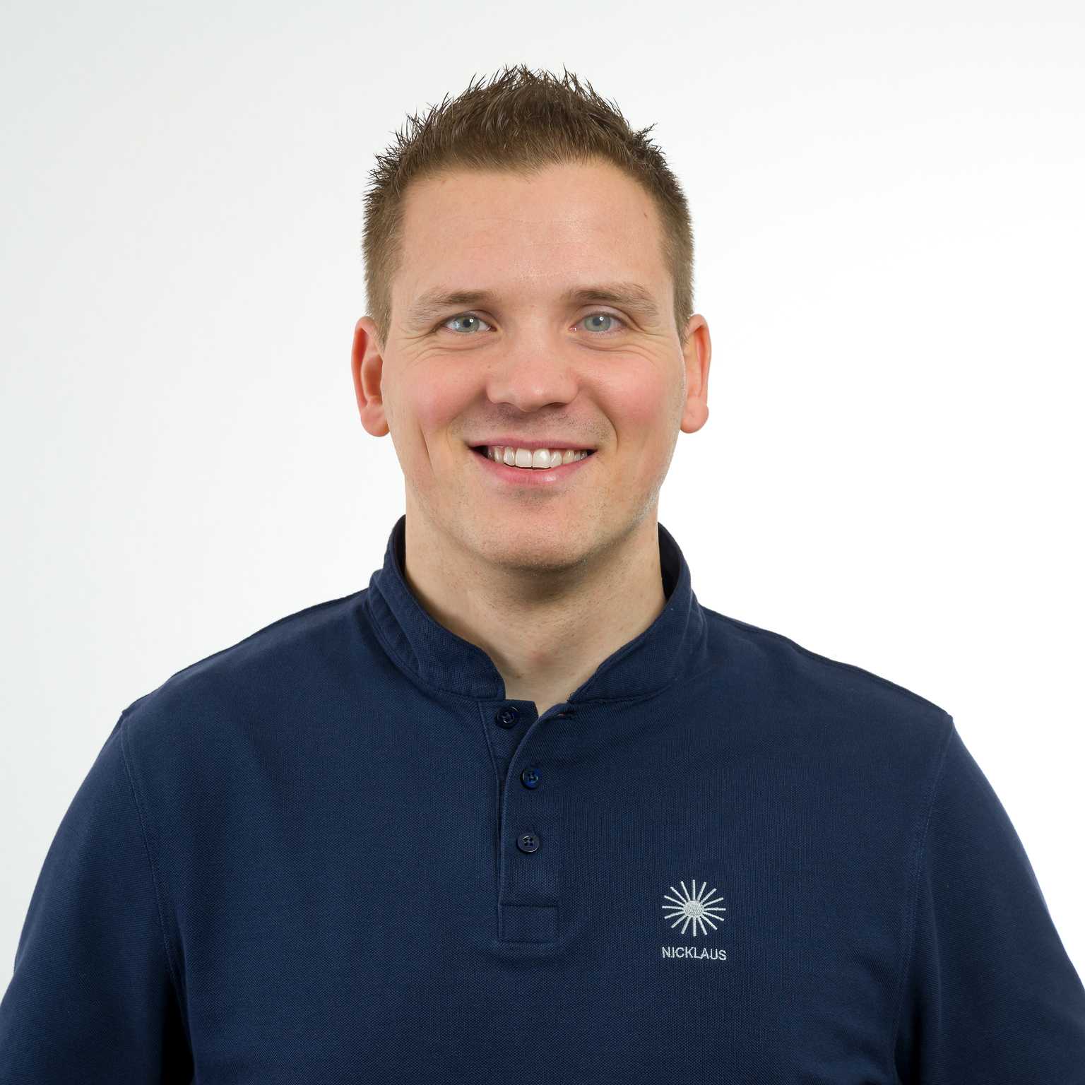 Portr&auml;t eines l&auml;chelnden Mannes mit kurzem, straffem Haar, der ein dunkles Poloshirt tr&auml;gt, vor hellem Hintergrund.