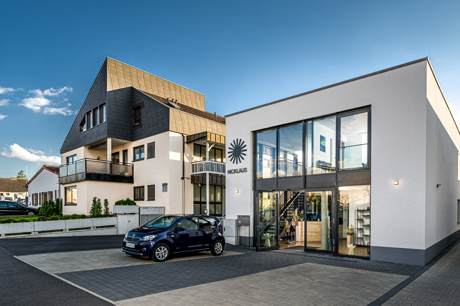 Moderne Geb&auml;ude mit Glasfassade und Treppe, davor ein blauer Smart und eine gepflegte Landschaft im Hintergrund.