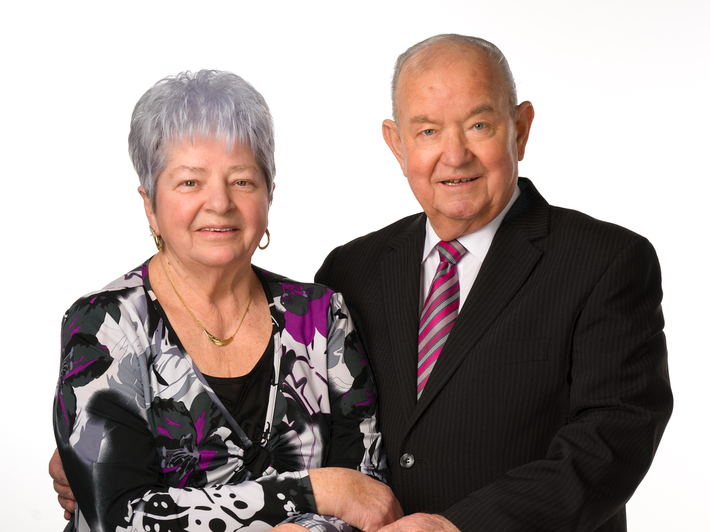 Ein &auml;lteres Paar posiert gemeinsam, l&auml;chelnd, vor einem hellen Hintergrund. Die Frau tr&auml;gt ein bl&uuml;hendes Oberteil, der Mann ein Nadelstreifenanzug.