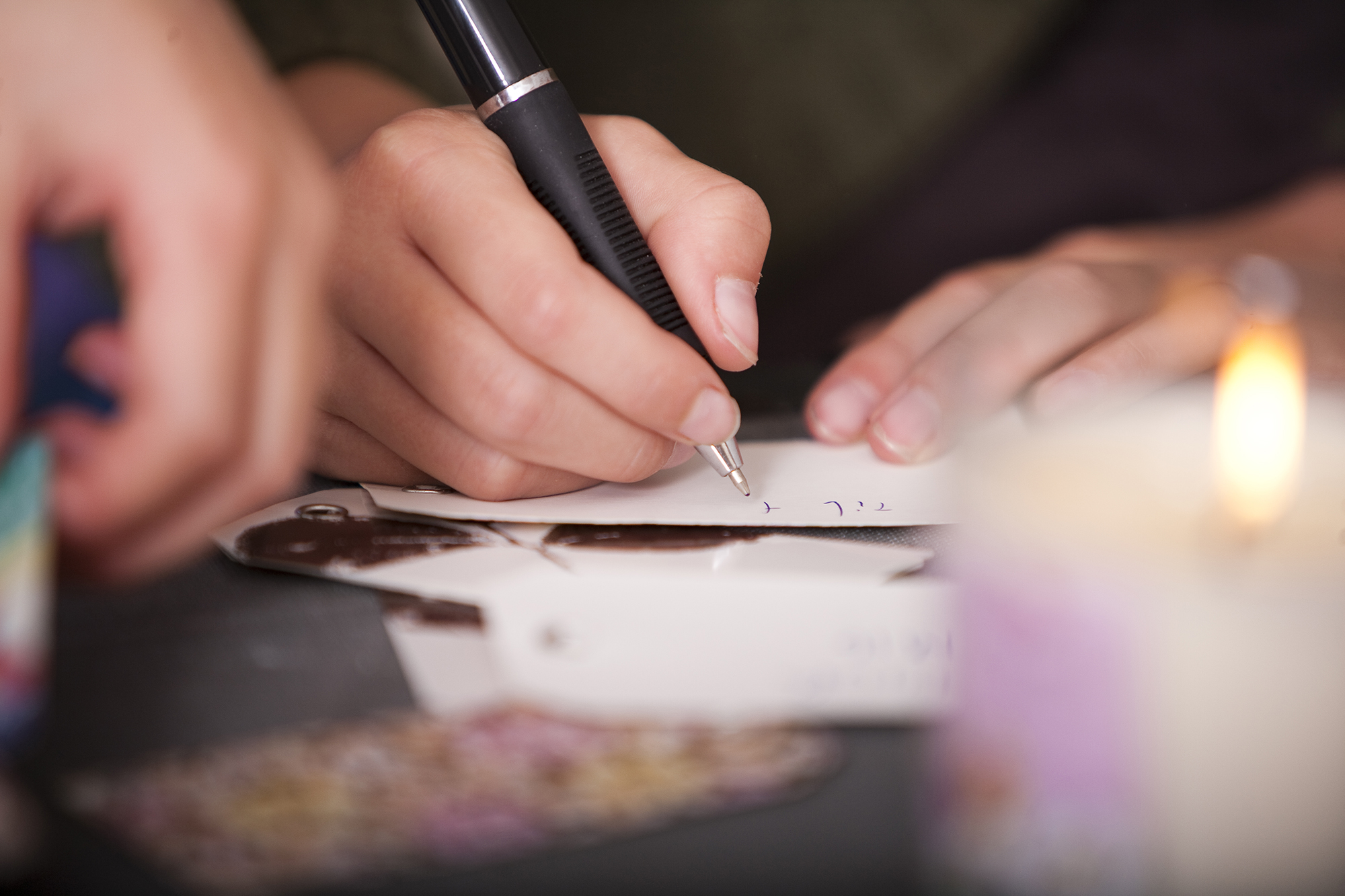 Nahaufnahme einer Hand, die mit einem Kugelschreiber auf einem Zettel schreibt, im Hintergrund eine brennende Kerze.