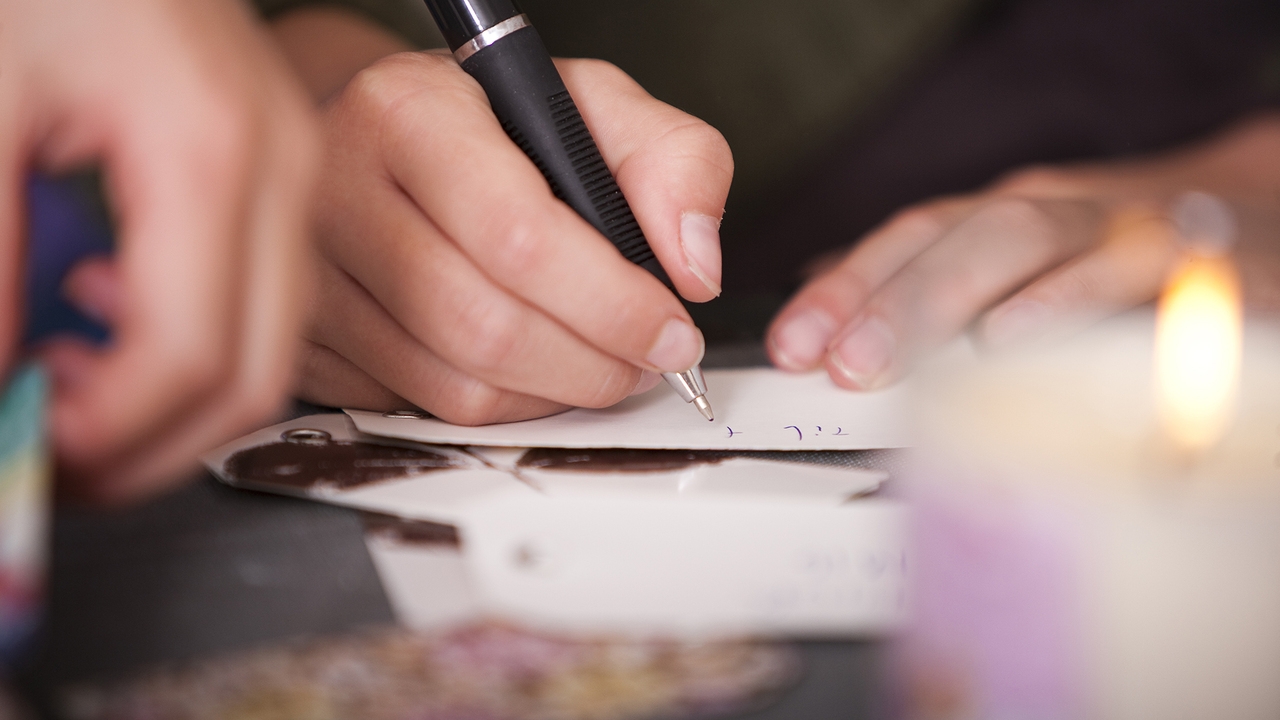 Nahaufnahme einer Hand, die mit einem Kugelschreiber auf einem Zettel schreibt, im Hintergrund eine brennende Kerze.