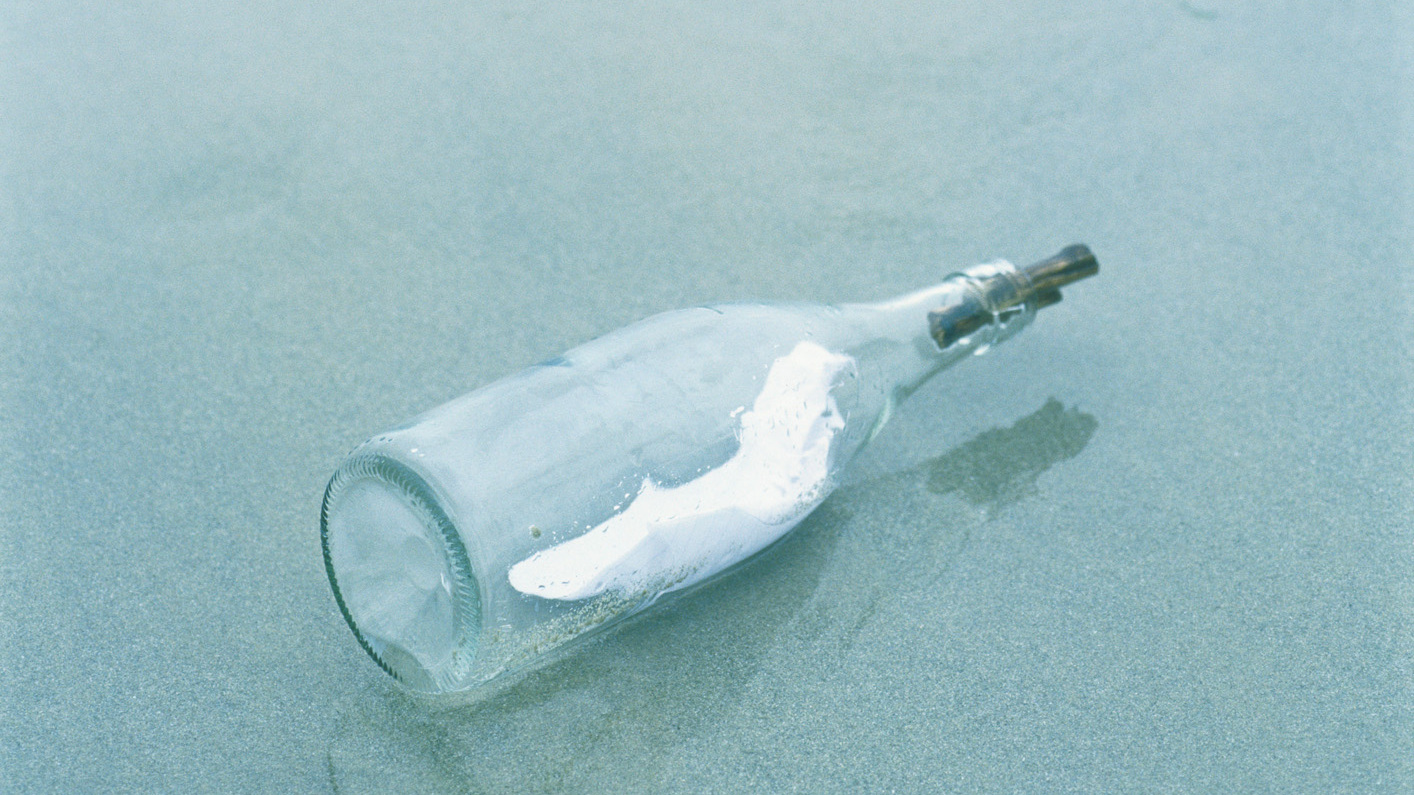 Eine Glasflasche liegt am Strand, umgeben von sanften Wellen und feinem Sand. In der Flasche ist ein dünnes Papier.